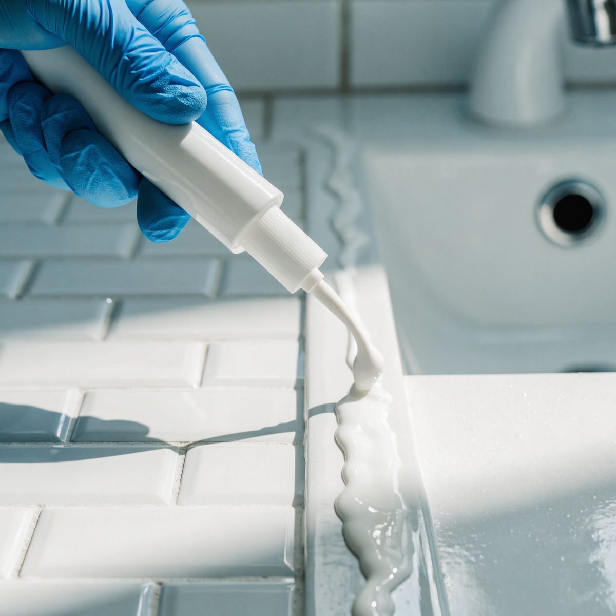 Gloved hand piping a thick bead of gel along a bathroom caulk line